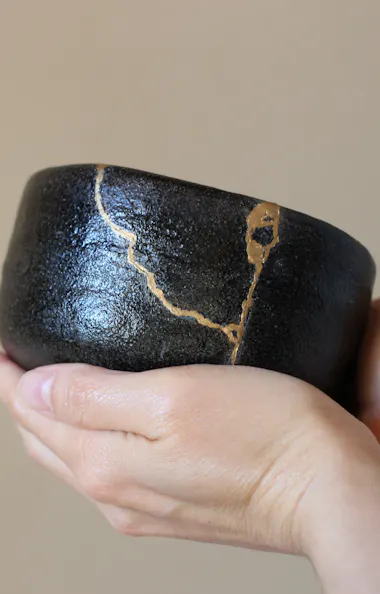 Kintsugi A person gently holds a black ceramic bowl repaired with visible gold seams, showcasing the Japanese art of Kintsugi against a neutral background.
