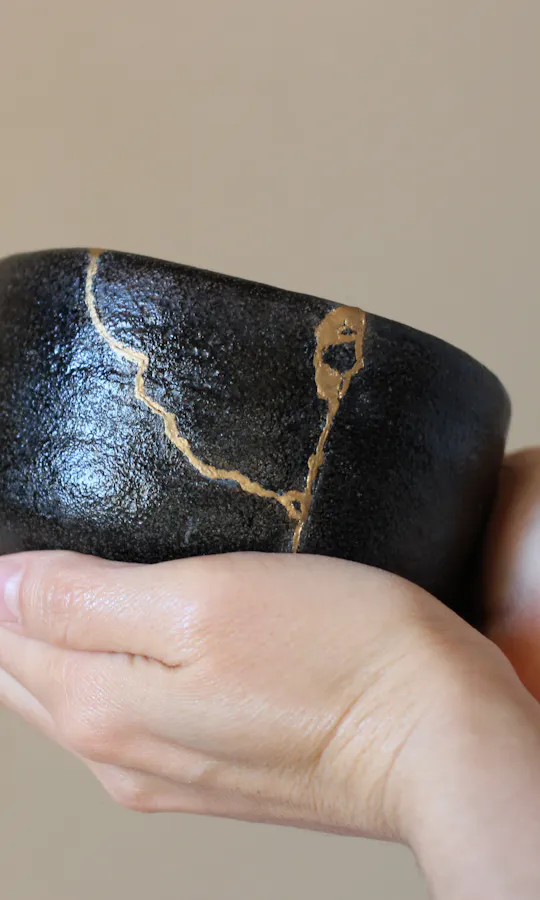 Kintsugi A person gently holds a black ceramic bowl repaired with visible gold seams, showcasing the Japanese art of Kintsugi against a neutral background.