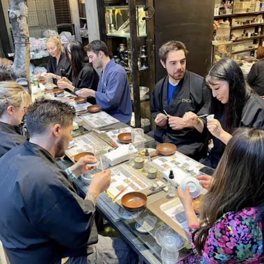 Kintsugi Workshop A group of people sit around a table in a pottery studio, wearing dark aprons and working on crafting or painting ceramic pieces. Shelves filled with pottery supplies and finished items line the background.
