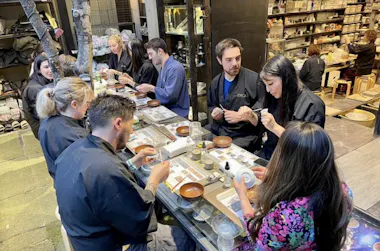 A group of people sit around a table in a pottery studio, wearing dark aprons and working on crafting or painting ceramic pieces. Shelves filled with pottery supplies and finished items line the background.
