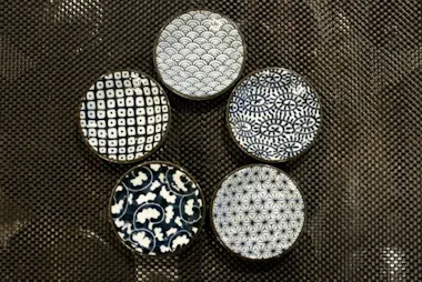 Five small round dishes with different blue and white geometric and floral patterns are arranged in a circle on a textured dark brown surface.