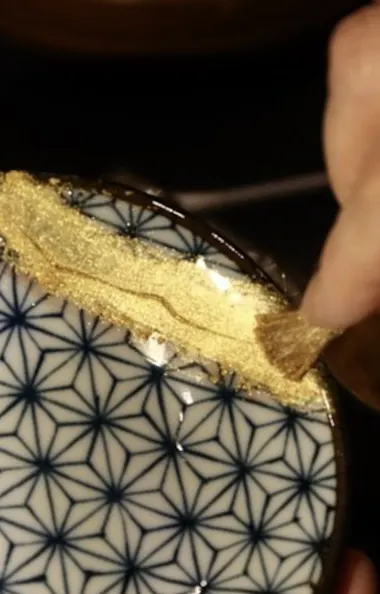Kintsugi Workshop A hand applies gold powder to a cracked ceramic bowl with a blue geometric pattern, repairing it using the Japanese Kintsugi technique.