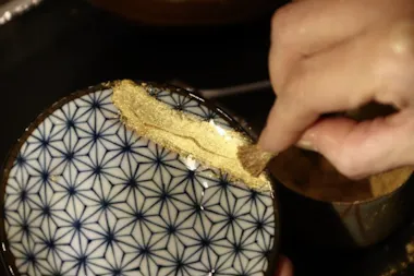 A hand applies gold powder to a cracked ceramic bowl with a blue geometric pattern, repairing it using the Japanese Kintsugi technique.