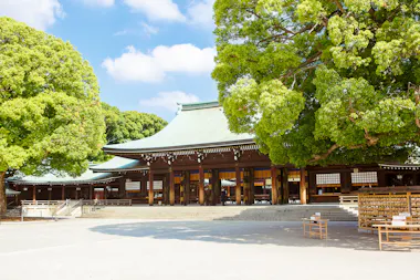 A traditional Japanese shrine with a green-tiled roof is surrounded by lush green trees under a blue sky. Wooden prayer plaques and benches are visible in the open courtyard.