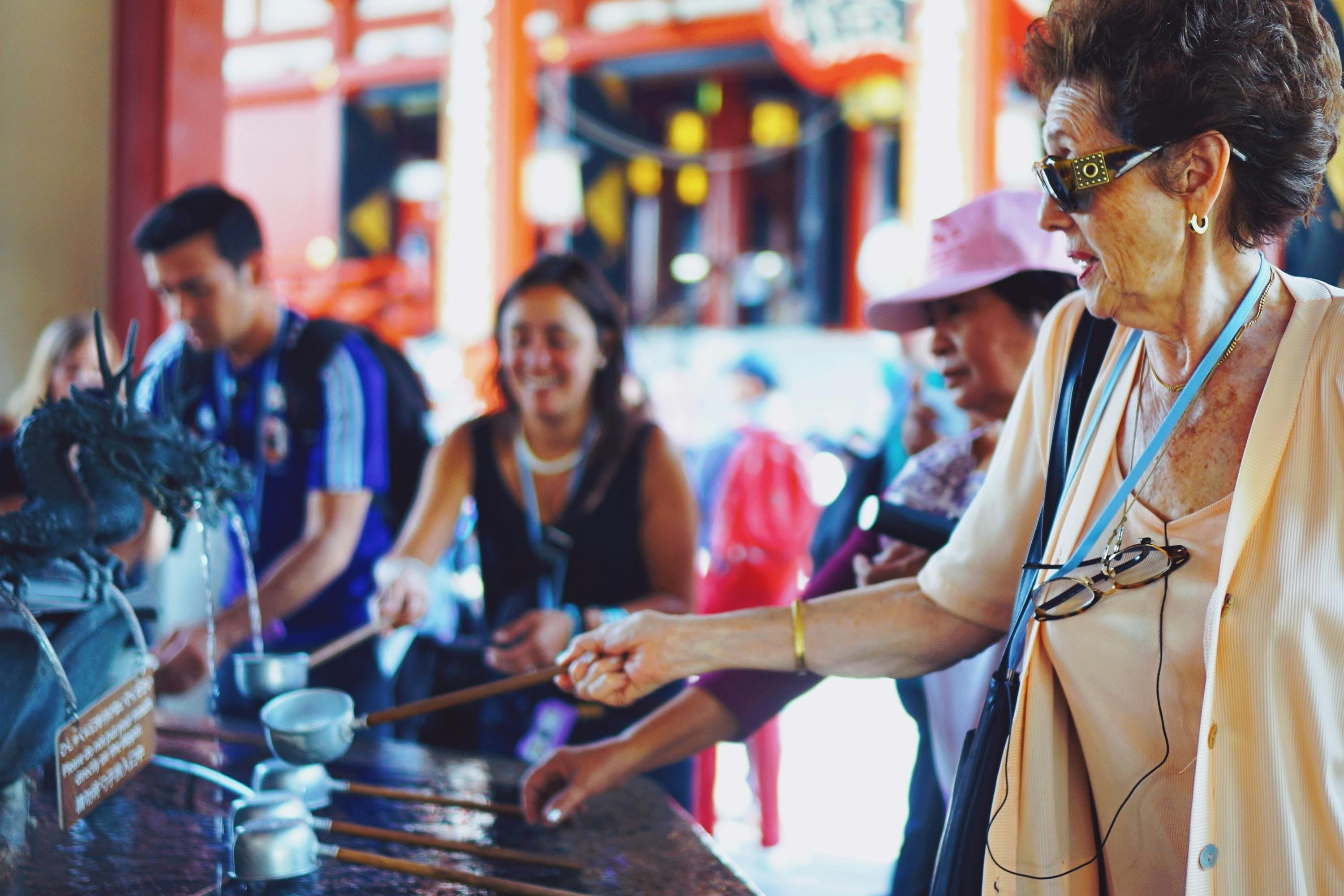 A group of people, including an elderly woman in sunglasses, use ladles to scoop water at a traditional temple purification fountain, with a dragon statue and others in the background.