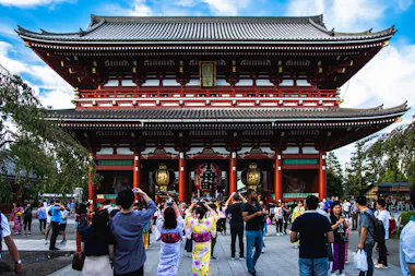 A large crowd gathers in front of Senso-ji Temple’s grand red entrance gate in Tokyo, Japan. Some people wear colorful yukatas and take photos, while others walk and admire the traditional architecture under a blue sky.