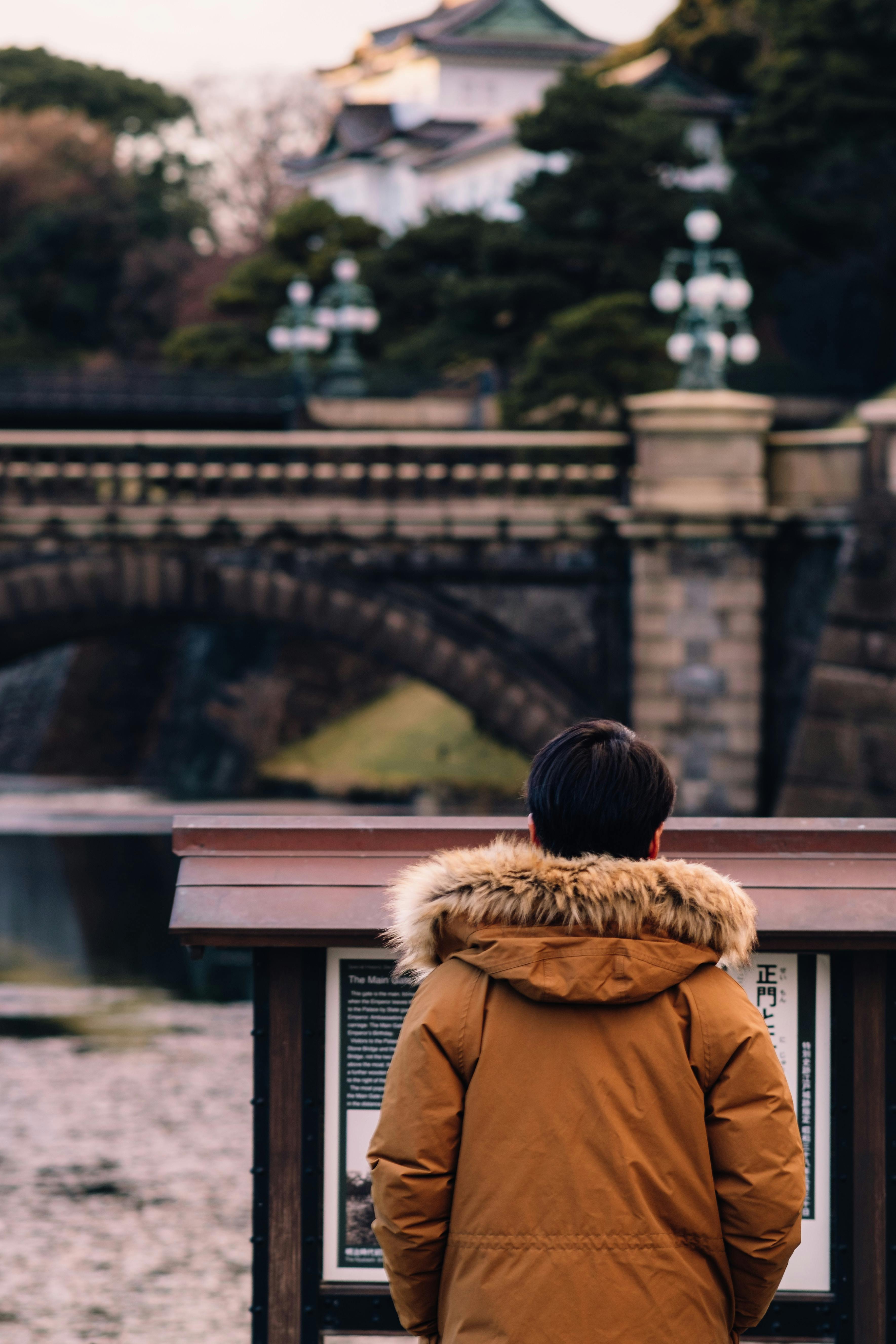 A person wearing a brown parka with a fur-lined hood stands in front of an information board, facing an old stone bridge and historic building surrounded by trees.