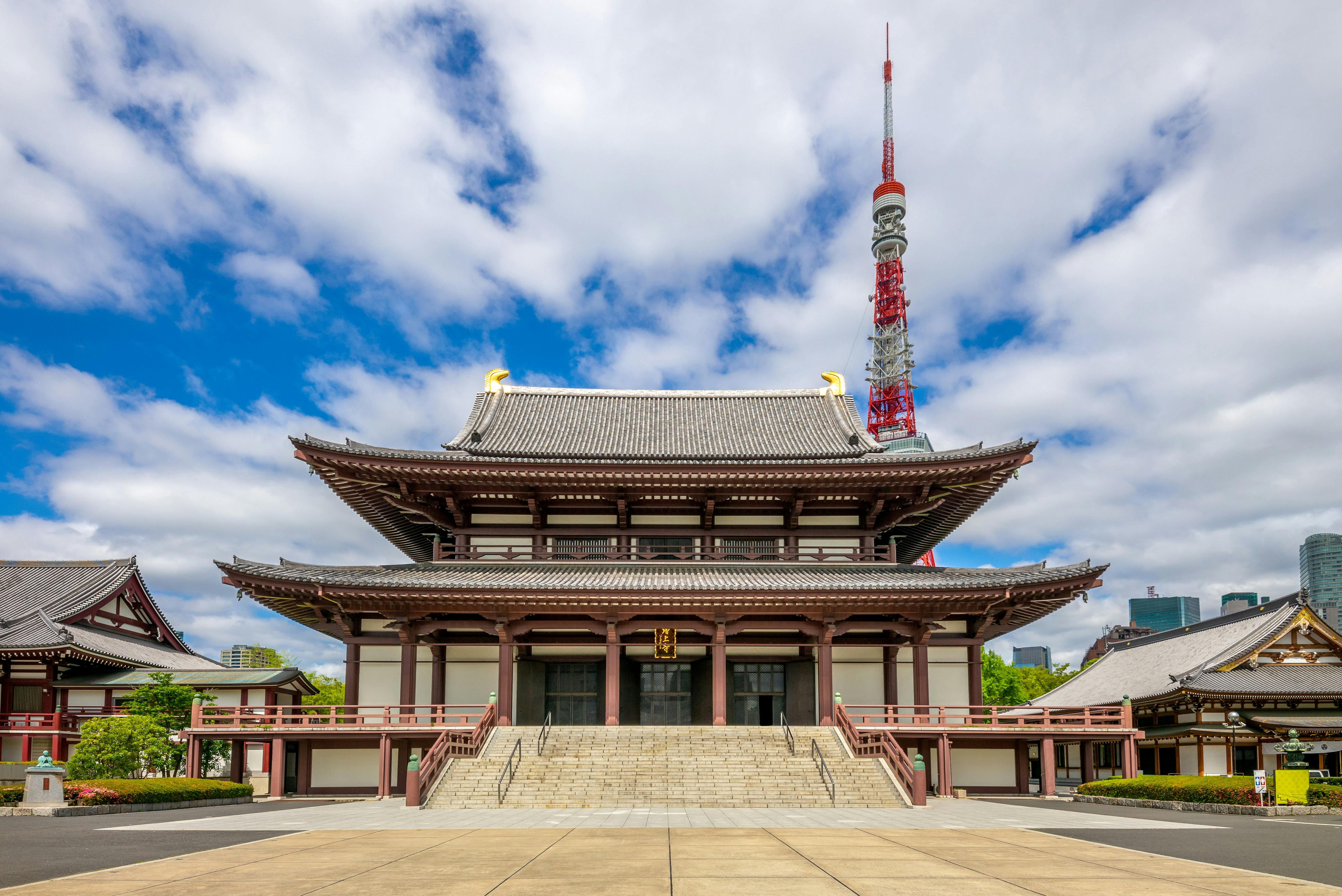 A traditional Japanese temple with a wide staircase and ornate roof stands in front of Tokyo Tower, under a partly cloudy sky, with modern buildings visible in the distant background.