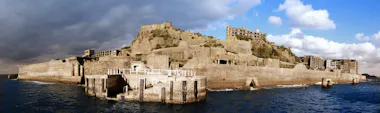 A panoramic view of an abandoned concrete island fortress rises from the sea, with crumbling buildings and walls under a partly cloudy sky.