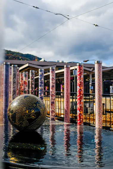 A black sphere with gold dragon designs sits on a reflective water surface, in front of colorful, patterned cylindrical pillars and a metal fence at a train station, with cloudy skies and hills in the background.