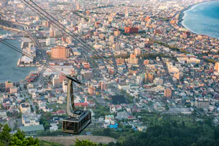 A cable car ascends above a coastal city with densely packed buildings, a harbor, and a curving shoreline next to the blue sea, viewed from an elevated vantage point.