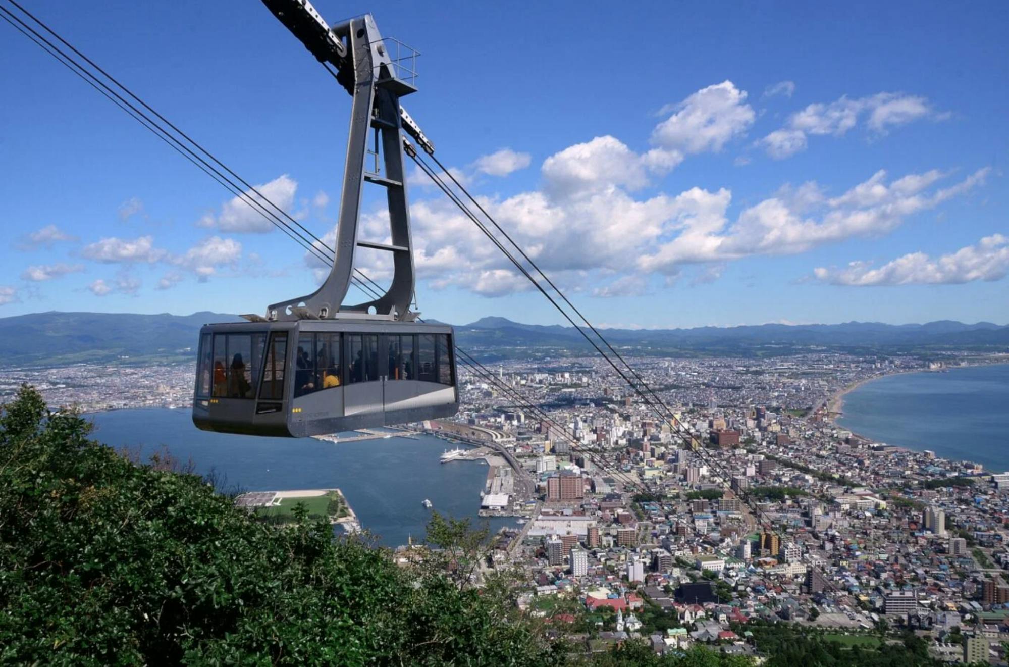 A cable car travels above a city with a harbor, with the coastline and buildings visible below, surrounded by hills and a blue sky with clouds.