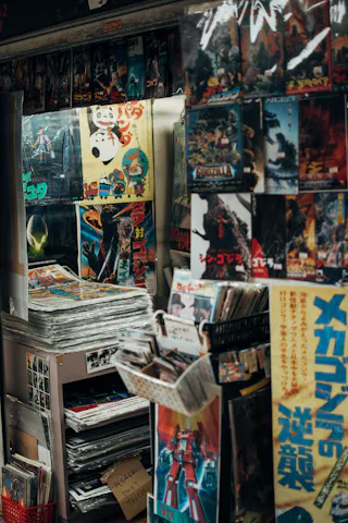 A shop display packed with colorful vintage Japanese posters, magazines, and comic books featuring various robots, monsters, and anime characters, with stacks of paper and shelves filled with more items.