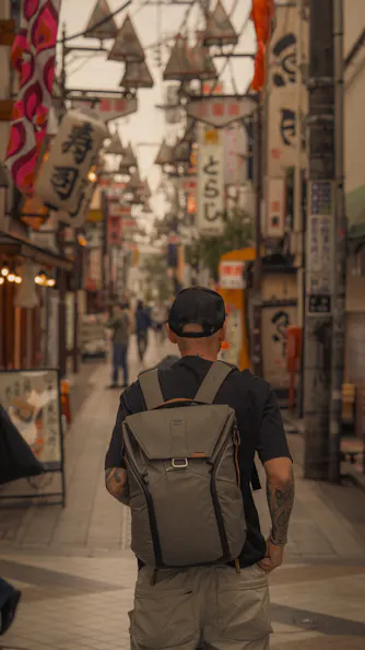 A person with tattoos, wearing a black cap and a gray backpack, walks down a narrow street lined with colorful signs and lanterns in what appears to be a Japanese city.