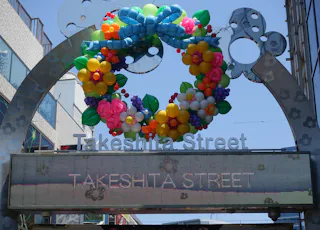 Entrance sign to Takeshita Street, decorated with a colorful, balloon floral wreath and a blue ribbon, against a clear blue sky. The digital marquee below also reads "Takeshita Street.