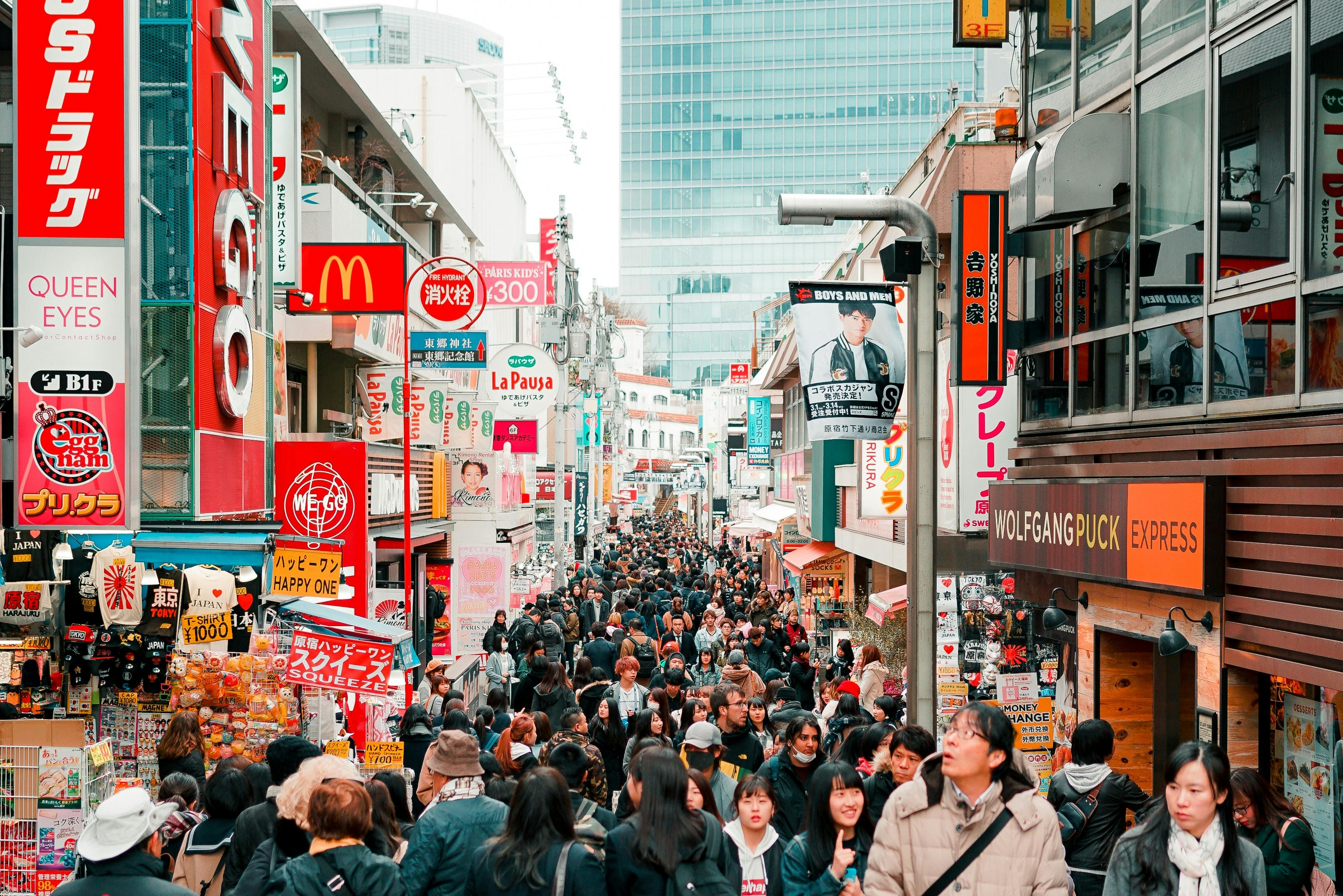 A busy city street crowded with people, surrounded by colorful shop signs and advertisements in Japanese. Buildings line both sides, and a McDonald's sign is visible among other shops.