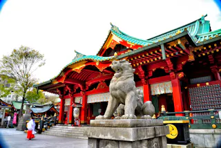 A traditional Japanese shrine with red columns, ornate green roofs, and a large stone guardian lion-dog statue in front. A person in white and red traditional attire walks nearby. Trees and lanterns are visible around the shrine.