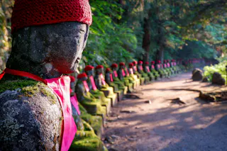 A row of moss-covered stone statues, each dressed in red hats and bibs, lines a sunlit forest path. The focus is on the nearest statue, with others fading into the distance.