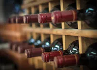 Close-up of several bottles of red wine stored horizontally in a wooden wine rack, with their red foil tops facing outward. The image is softly focused, highlighting the bottles and rack rows.