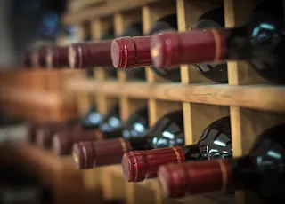 Close-up of several bottles of red wine stored horizontally in a wooden wine rack, with their red foil tops facing outward. The image is softly focused, highlighting the bottles and rack rows.