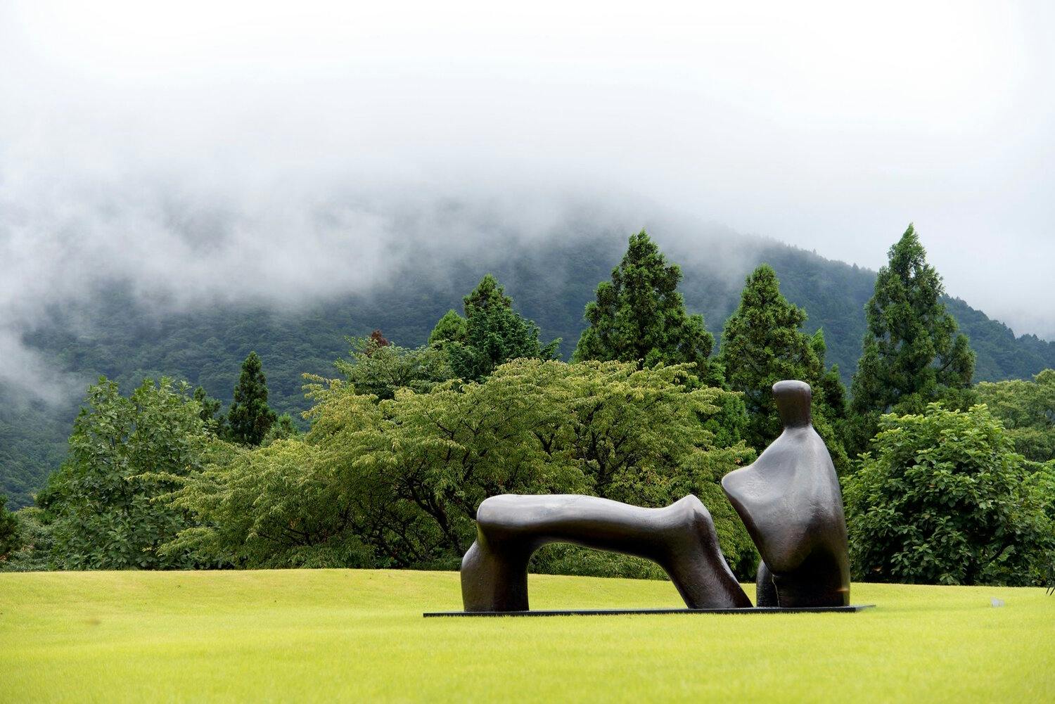 A modern abstract sculpture stands in a lush, green field surrounded by thick, tall trees. In the background, mist and clouds partially obscure forested hills and mountains, adding a mysterious and serene ambiance to the landscape.