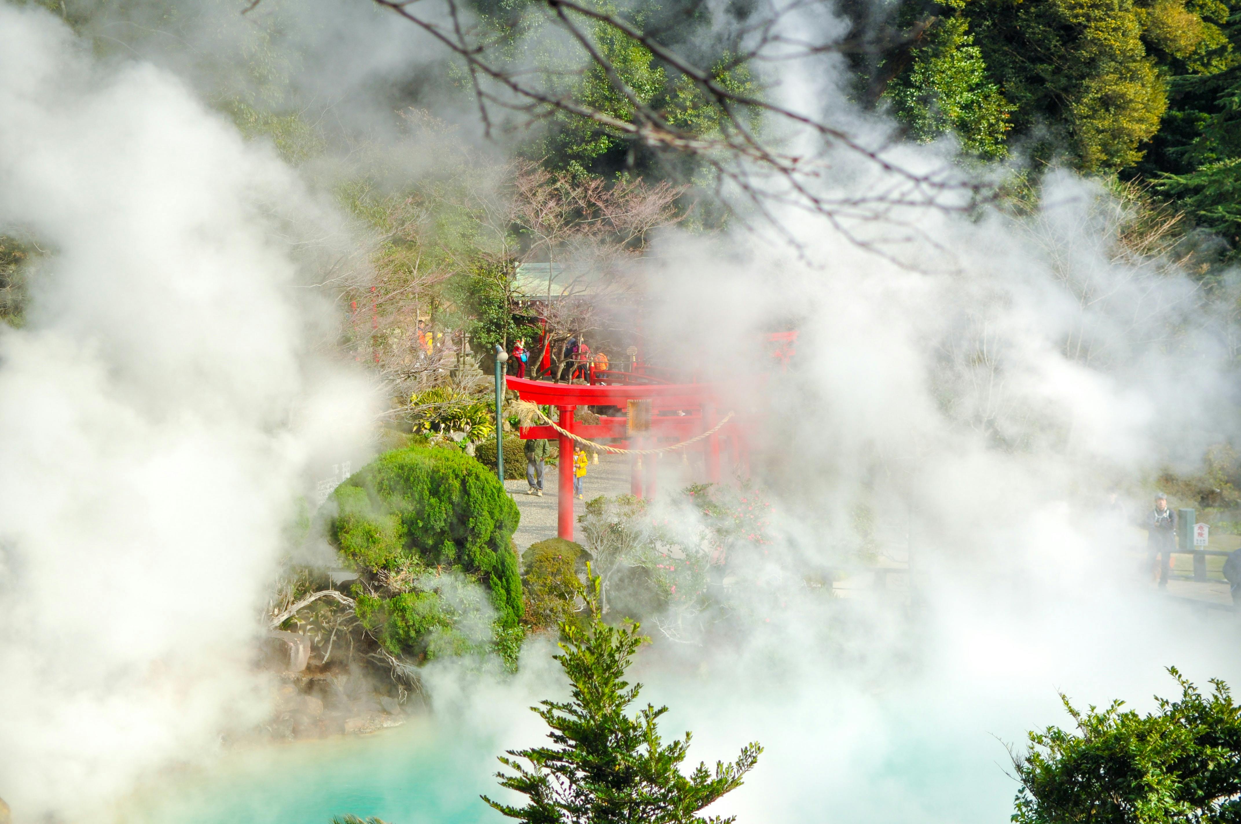 Steam rises from a hot spring surrounded by lush greenery and trees, partially obscuring a vibrant red torii gate in a Japanese garden setting.