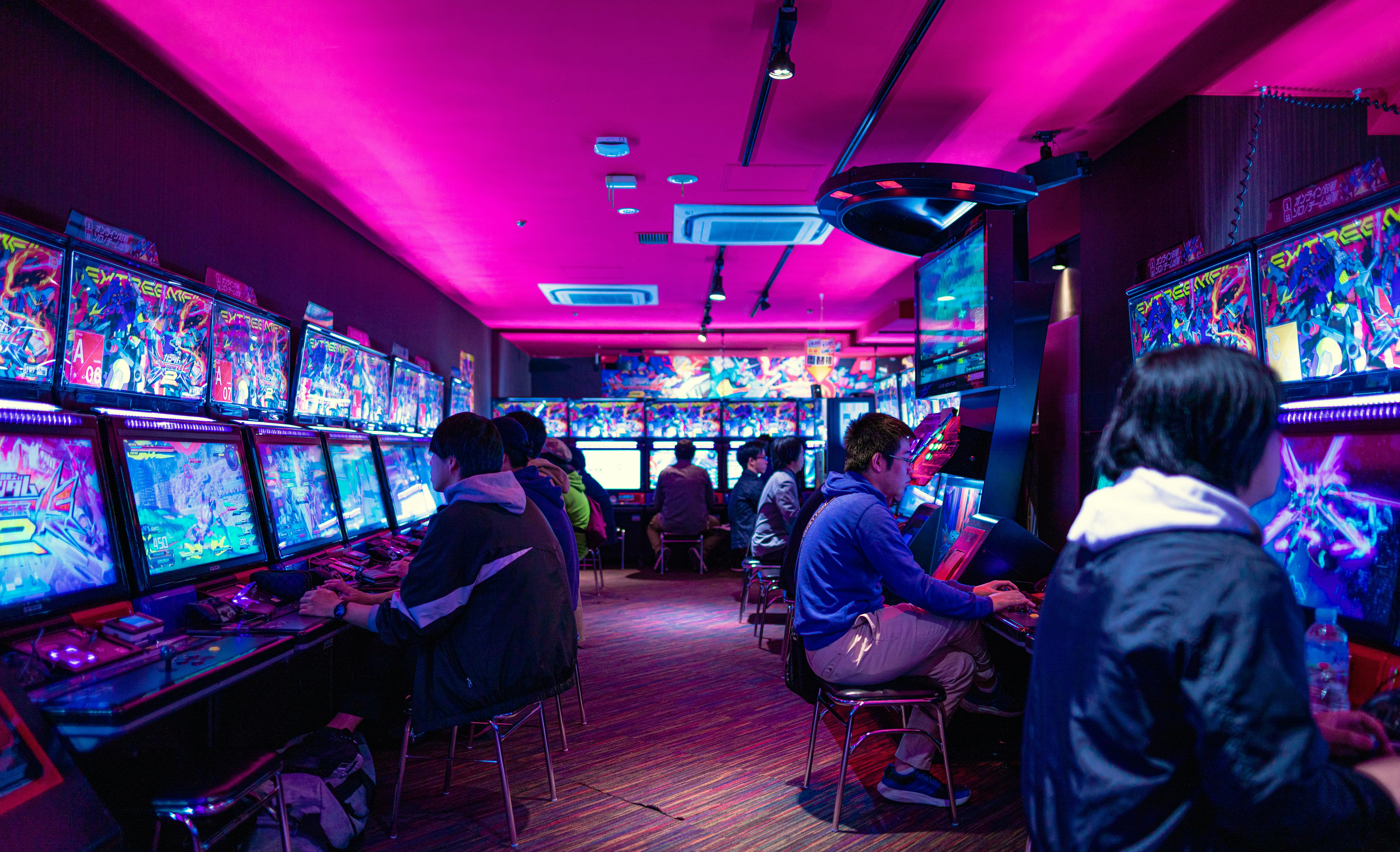 People sit at arcade game machines in a neon-lit room with vibrant pink lighting, playing video games. The arcade is busy, with many screens displaying colorful graphics and several people wearing jackets and hoodies.