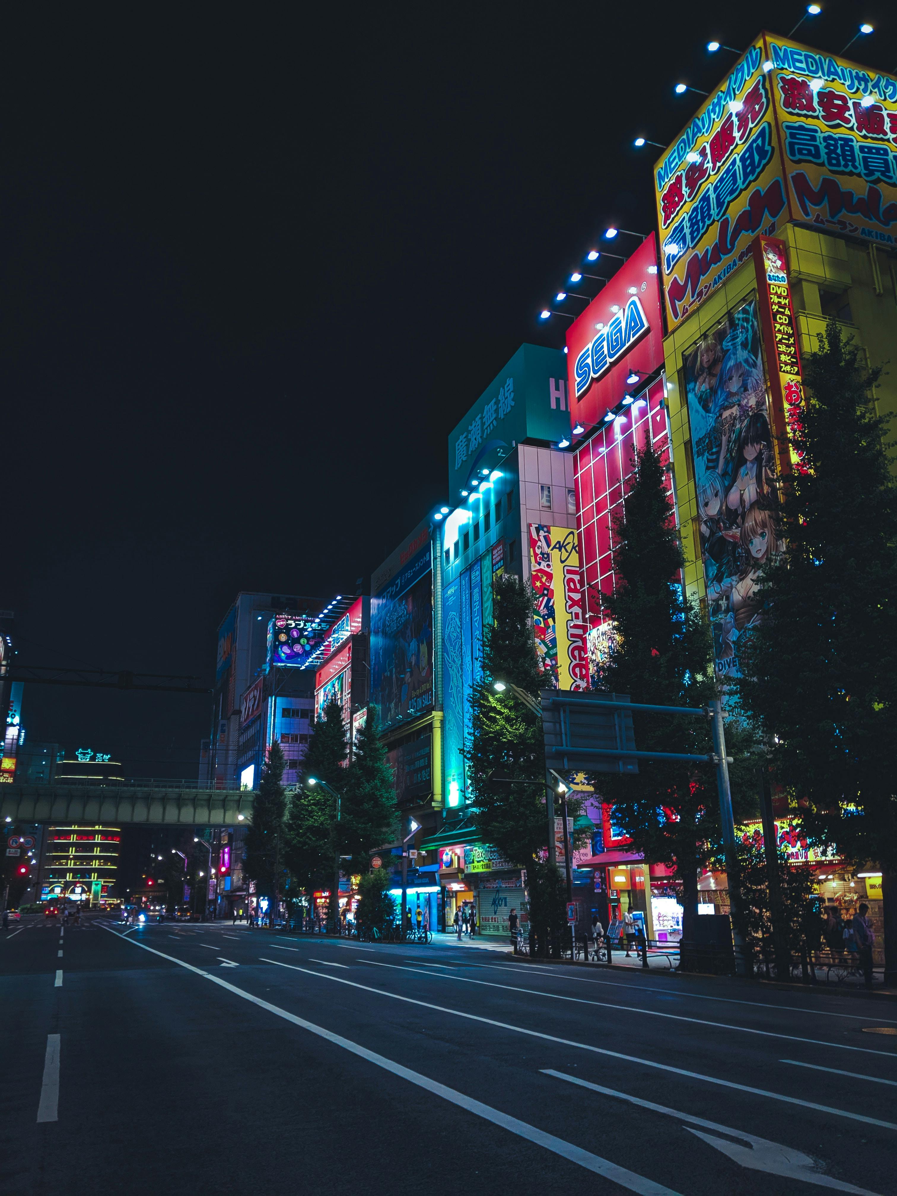 A city street at night lined with tall buildings covered in colorful, glowing neon signs in Japanese, with anime and game advertisements, and very few people or cars visible.