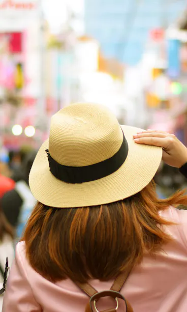 Takeshita Street A person wearing a beige hat and pink coat stands in a busy street market, surrounded by blurred crowds and colorful signs, seen from behind on a bright day.