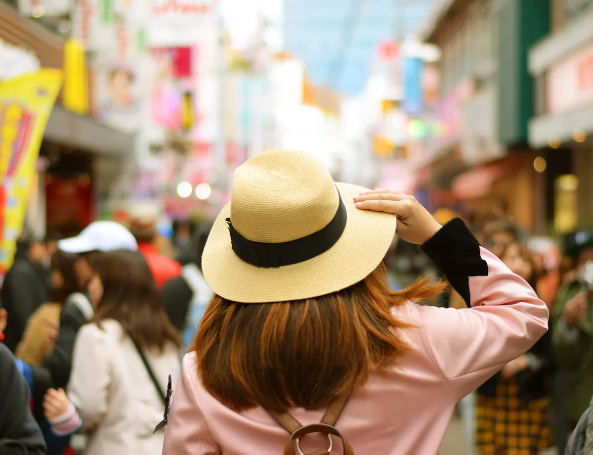Takeshita Street A person wearing a beige hat and pink coat stands in a busy street market, surrounded by blurred crowds and colorful signs, seen from behind on a bright day.