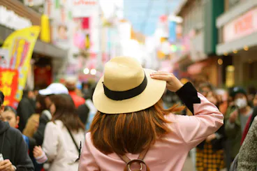 A person wearing a beige hat and pink coat stands in a busy street market, surrounded by blurred crowds and colorful signs, seen from behind on a bright day.