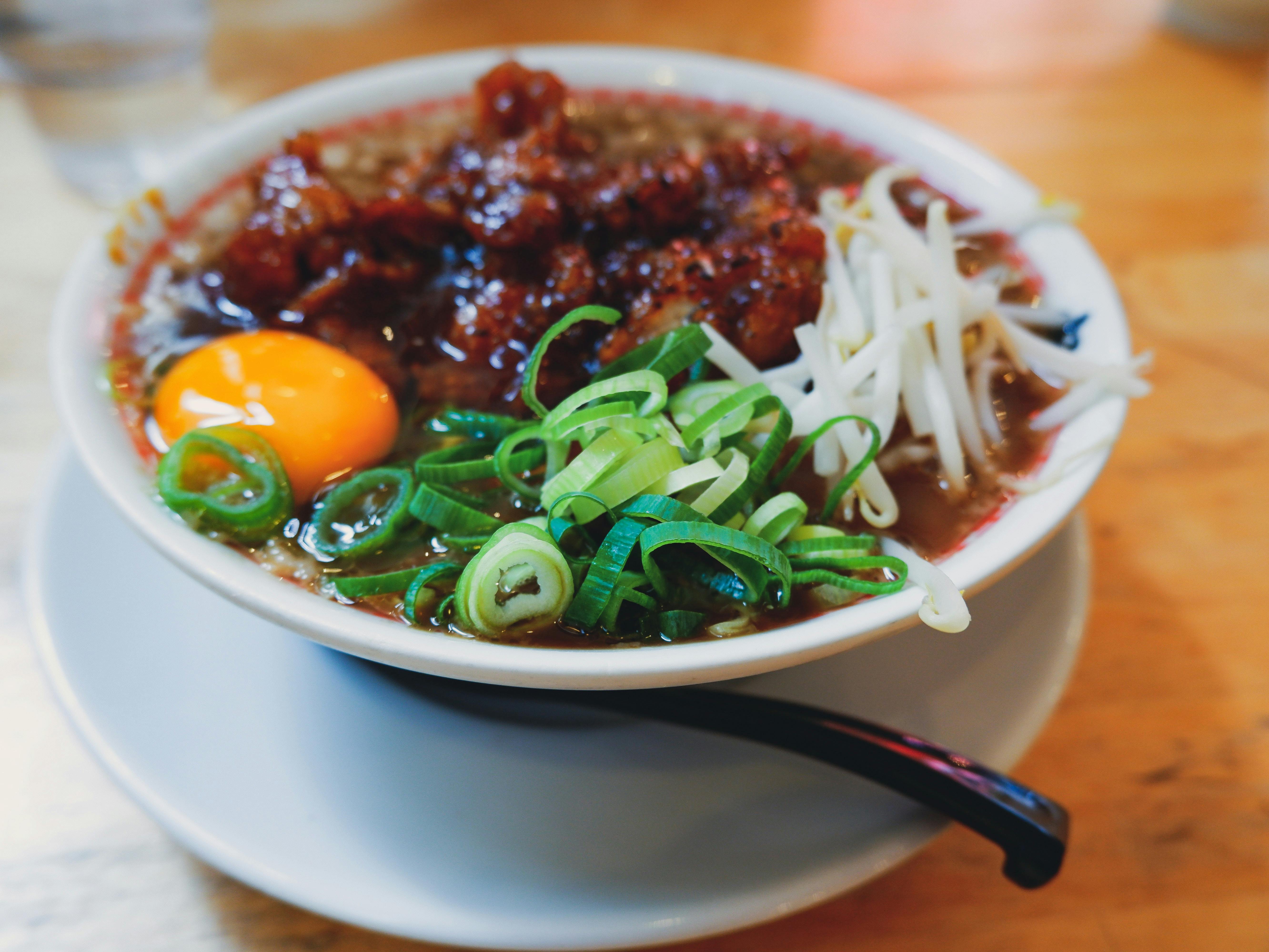 A bowl of ramen topped with a raw egg yolk, sliced green onions, bean sprouts, and pieces of crispy meat, served with a black spoon on a white plate.