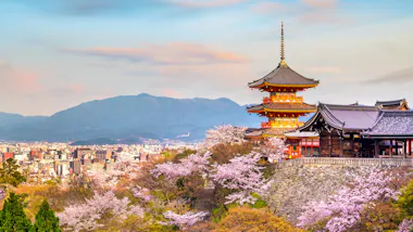 A traditional Japanese pagoda stands among cherry blossoms, overlooking a city with mountains in the background under a colorful sky.