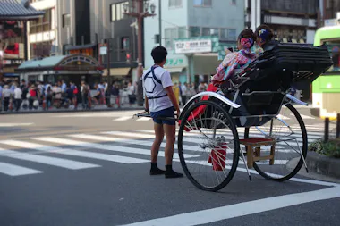 A rickshaw puller waits at a crosswalk with two women in traditional Japanese kimonos seated in the rickshaw. Urban buildings, a crowd, and a bus are visible in the background.