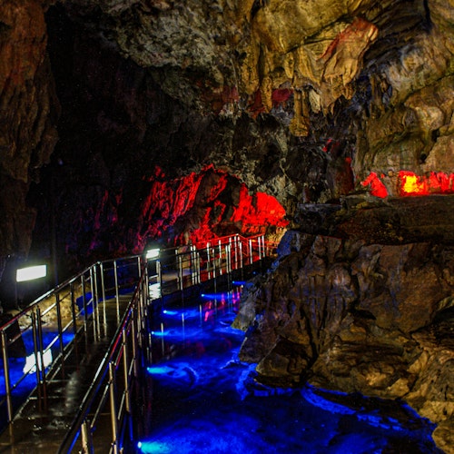 Hida Great Limestone Cave A winding metal walkway inside a cave is illuminated by vibrant blue and red lights, highlighting jagged rock walls and creating colorful reflections on the wet surface.