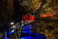 A winding metal walkway inside a cave is illuminated by vibrant blue and red lights, highlighting jagged rock walls and creating colorful reflections on the wet surface.