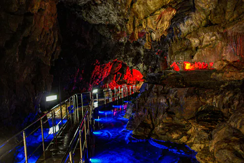 A winding metal walkway inside a cave is illuminated by vibrant blue and red lights, highlighting jagged rock walls and creating colorful reflections on the wet surface.