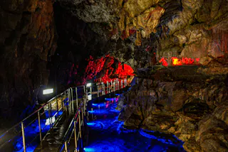 A winding metal walkway inside a cave is illuminated by vibrant blue and red lights, highlighting jagged rock walls and creating colorful reflections on the wet surface.