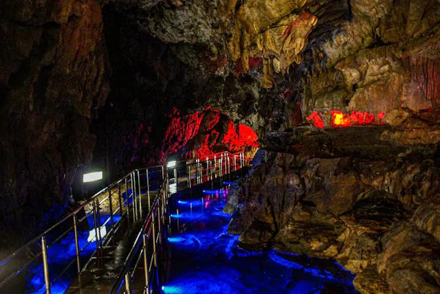 A winding metal walkway inside a cave is illuminated by vibrant blue and red lights, highlighting jagged rock walls and creating colorful reflections on the wet surface.