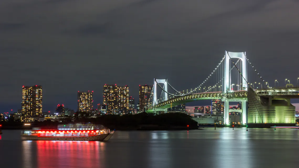 Rainbow Bridge at night Night view of a brightly lit bridge spanning a body of water, with a boat illuminated by red lights in the foreground. In the background, tall city buildings are also lit up, reflecting on the calm water. The sky is dark and cloudy.