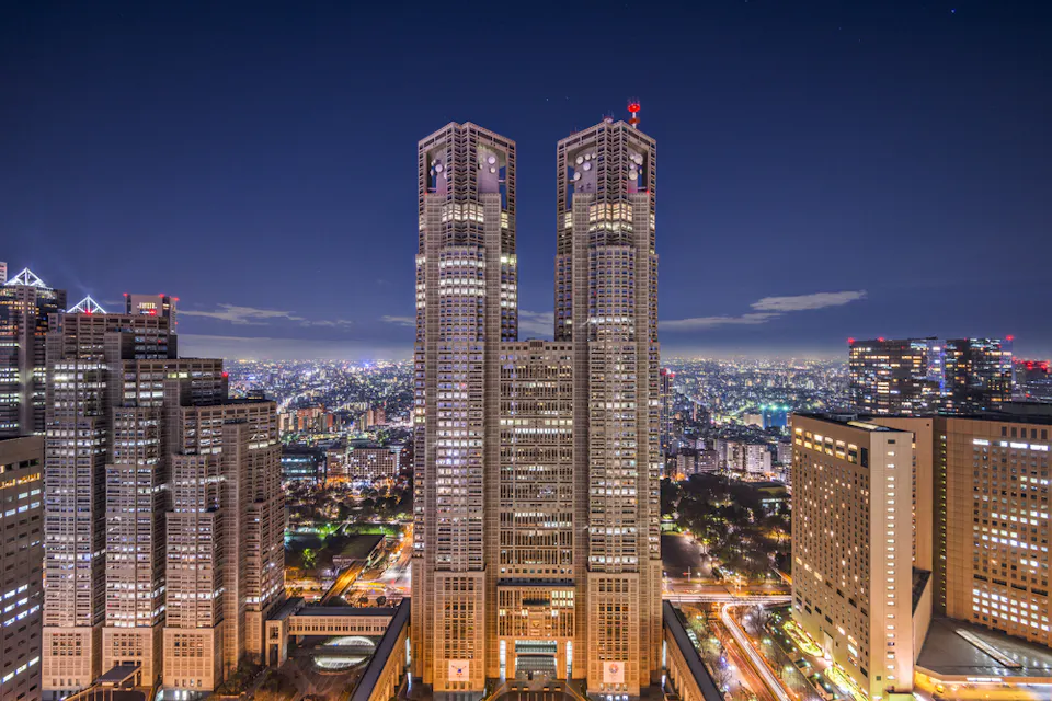 Tokyo Metropolitan Government Building at night A nighttime cityscape featuring a pair of tall, illuminated skyscrapers in the center, surrounded by other buildings. The sky is clear with a few scattered clouds, and the city lights create a vibrant, glowing scene.