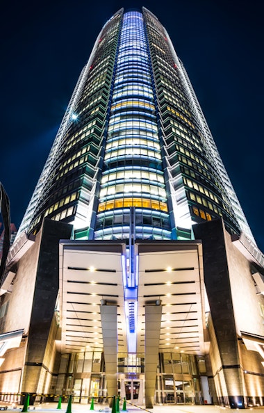 Roppongi Hills Mori Tower at Night A towering modern skyscraper is illuminated against the night sky, showcasing its sleek architecture. A large, abstract sculpture resembling a spider is visible to the left. The surrounding area is dotted with lights and additional buildings.