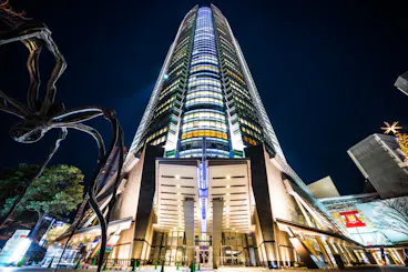 A towering modern skyscraper is illuminated against the night sky, showcasing its sleek architecture. A large, abstract sculpture resembling a spider is visible to the left. The surrounding area is dotted with lights and additional buildings.