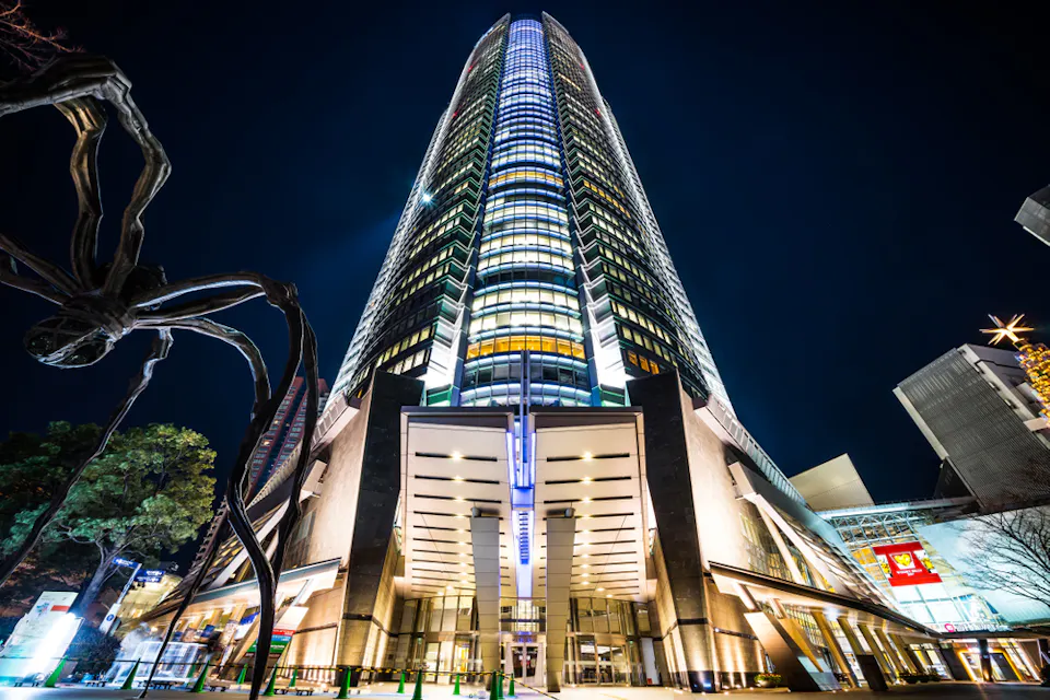 Roppongi Hills Mori Tower at Night A towering modern skyscraper is illuminated against the night sky, showcasing its sleek architecture. A large, abstract sculpture resembling a spider is visible to the left. The surrounding area is dotted with lights and additional buildings.