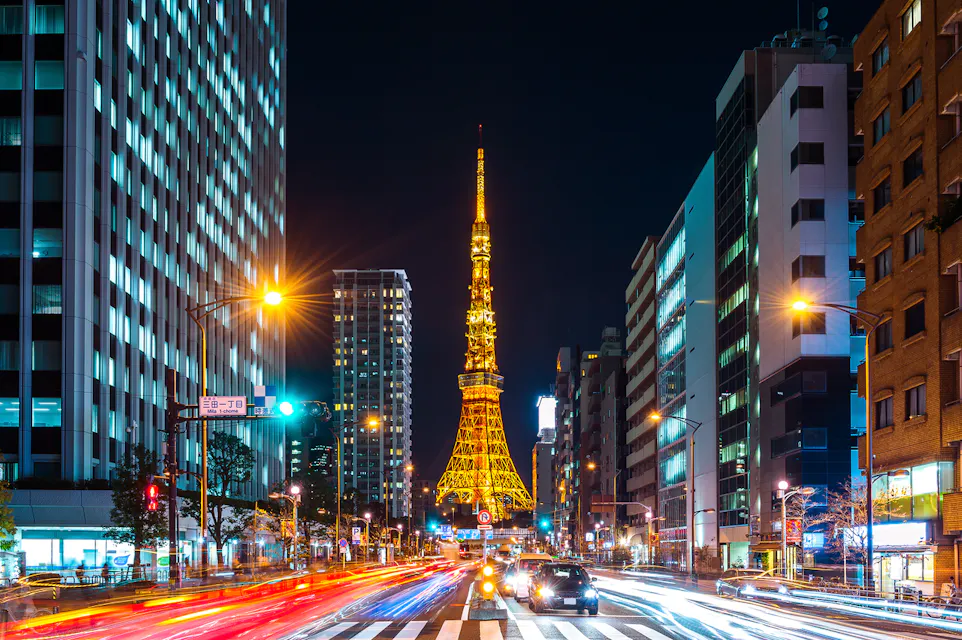 Tokyo Tower view at night A bustling city scene at night featuring Tokyo Tower illuminated in the center. Tall buildings line the street while car lights create dynamic streaks of red and white, adding energy to the vibrant urban landscape.