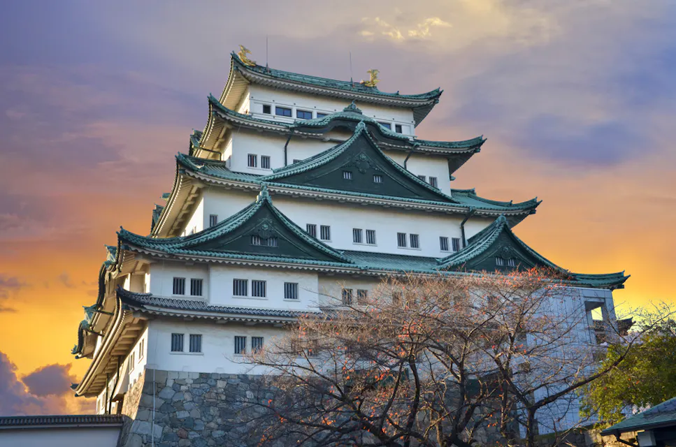 一座雄伟的城堡，带有多重阶梯屋顶和绿色装饰，显著地矗立在生动的日落天空下。传统的日本建筑被光秃的树木环绕，结构的石基部分可见。