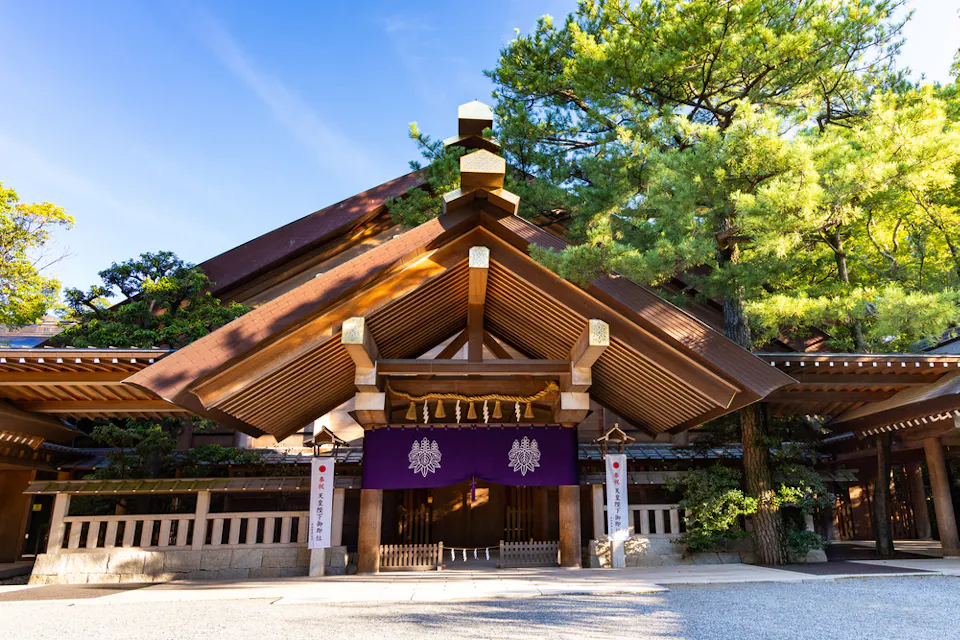 一座神社入口，特色为传统木结构的大屋檐，悬挂着装饰有白色标志的紫色幕帘。周围环绕着浓密绿叶的树木，神社沐浴在明媚的阳光下。一条碎石小路通向入口。