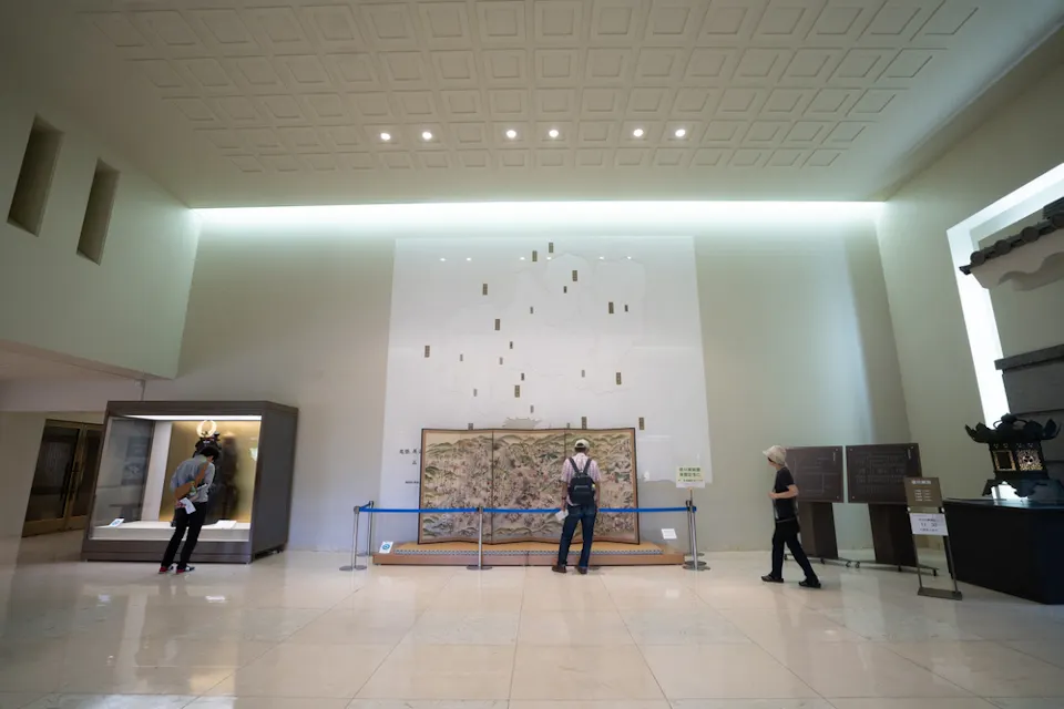 三个人正在一个拥有高天花板和极简设计的博物馆里观看展品。一个人正看着左侧的玻璃展示柜，另一个人正在中心查看一件大型艺术品，第三个人则站在房间的右侧。