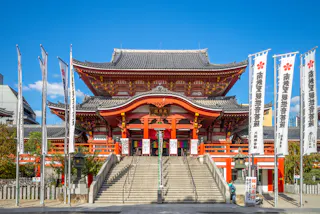 A large, traditional Japanese temple with ornate red and white details stands under a clear blue sky. Stone steps lead up to the entrance, flanked by tall banners with Japanese writing. Trees and additional buildings surround the temple grounds.