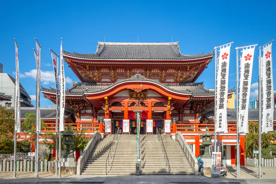一座大型传统日本寺庙，带有华丽的红白色细节，矗立在晴朗的蓝天之下。石阶通向入口，两旁是插有日文横幅的高旗。树木和其他建筑环绕在寺庙的院子周围。